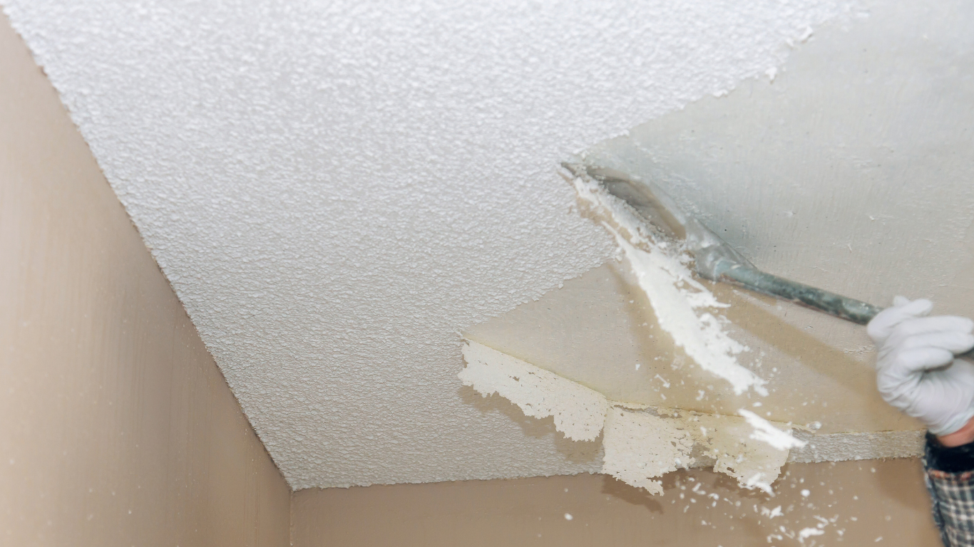 Person scraping textured ceiling, flaking off chunks with a scraper. Beige walls visible.