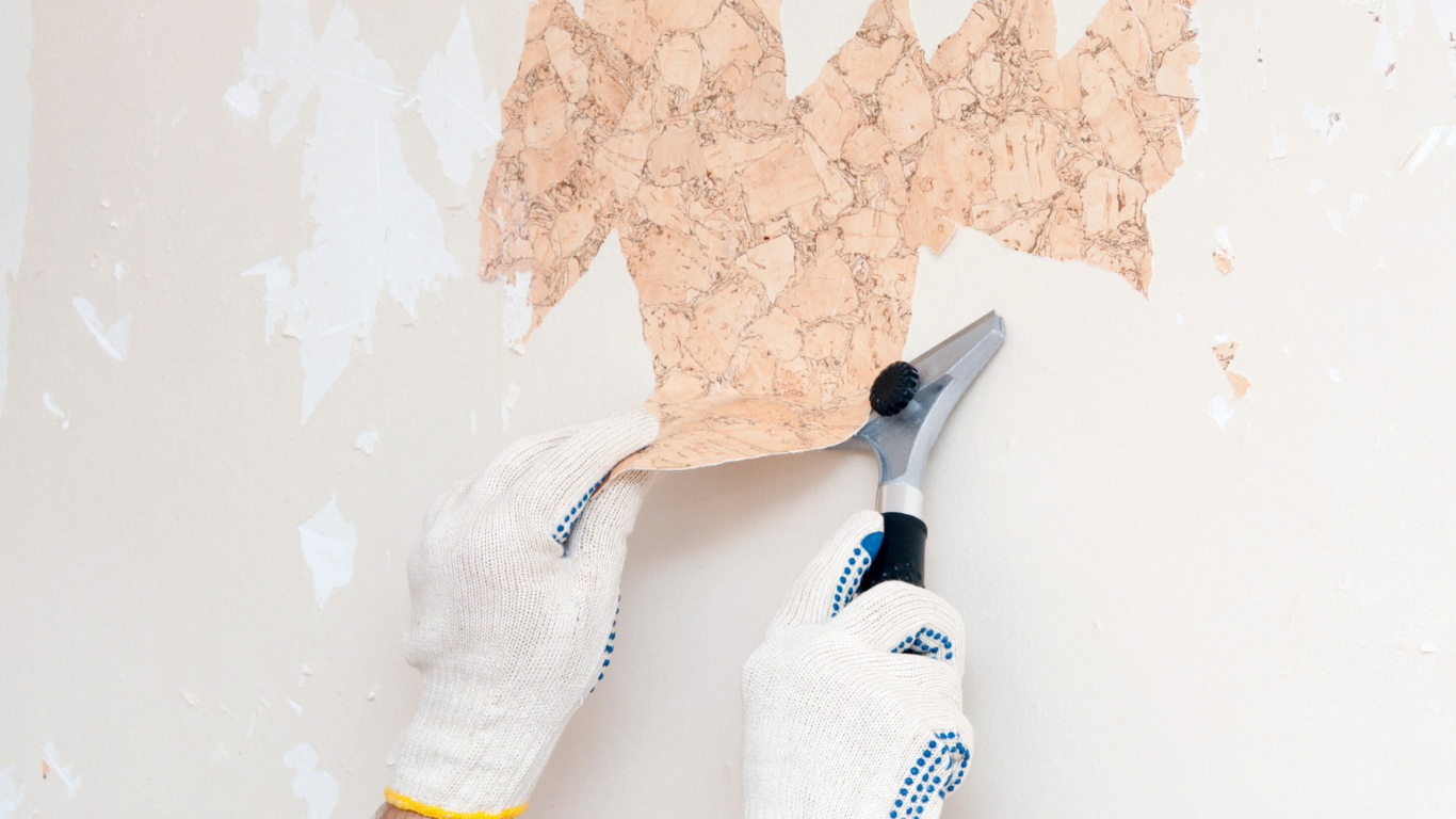 Hands in work gloves removing wallpaper with a scraper.
