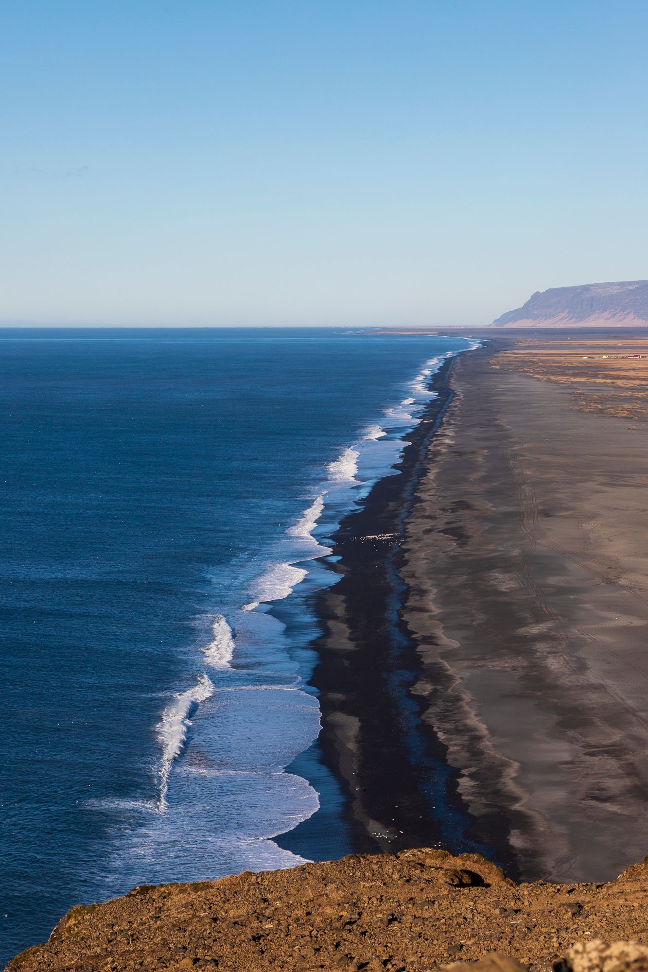 Een kustlijn met blauwe oceaangolven naast een donker rotsachtig strand en kliffen in de verte onder een heldere hemel.