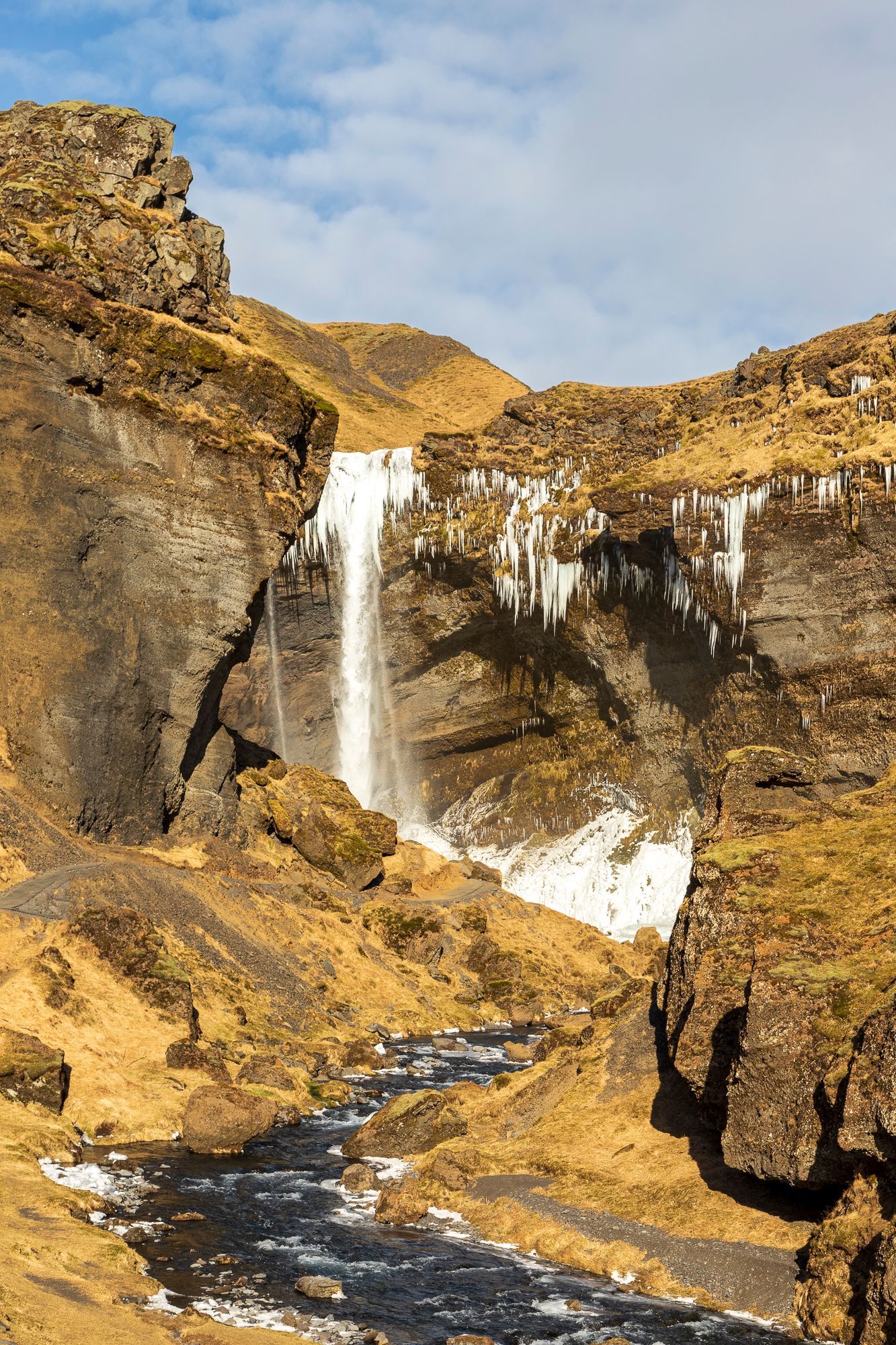 Een waterval stort neer in een rotsachtige kloof, met daaronder een besneeuwde beek onder een bewolkte hemel.