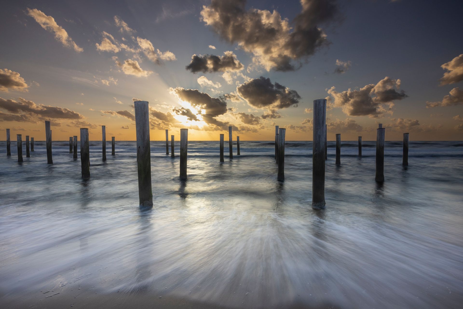 Zonsondergang boven een kalm strand met houten palen die uit het water oprijzen en dramatische wolken aan de hemel.