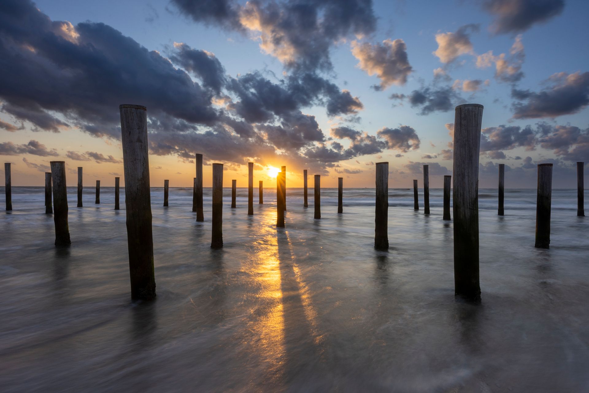 Zonsondergang boven ondiep water met houten palen en gloeiende reflecties onder dramatische wolken.