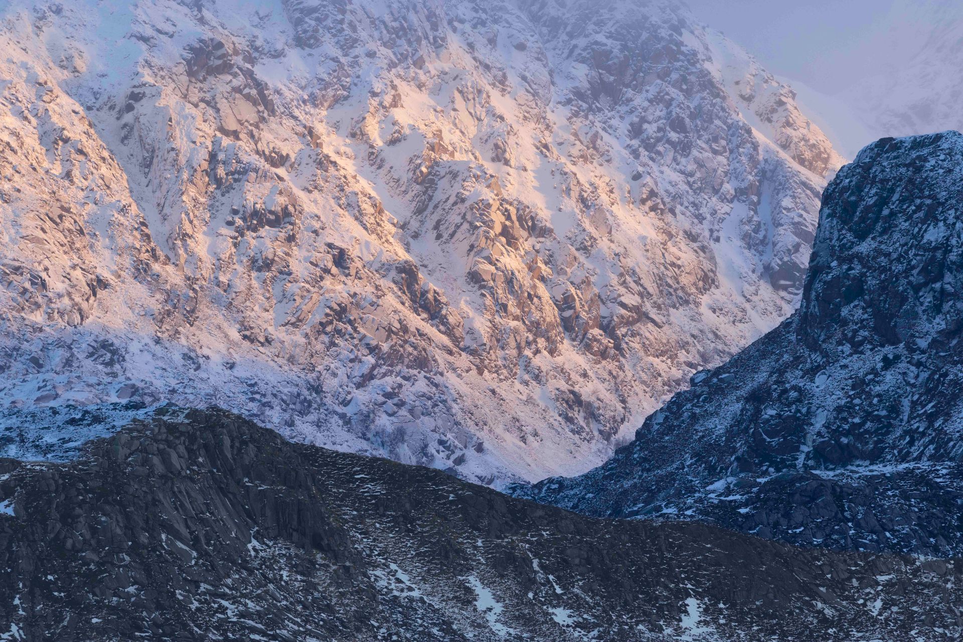 Een met sneeuw bedekte bergtop bij zonsopgang, die roze oplicht tegen een donkere, beboste bergkam.