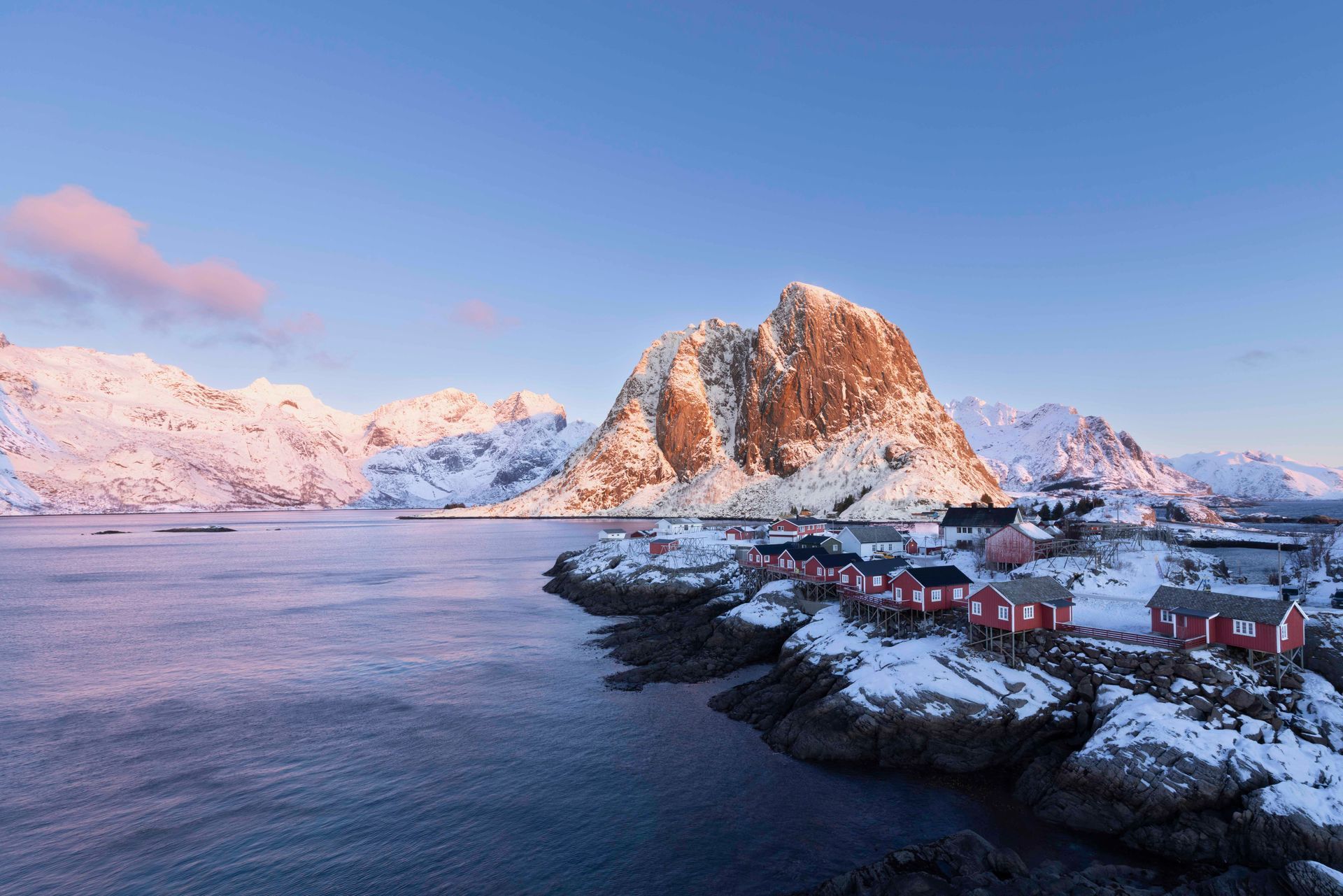 Een besneeuwd kustdorpje aan een kalme fjord met op de achtergrond een steile, roze verlichte berg bij zonsondergang.