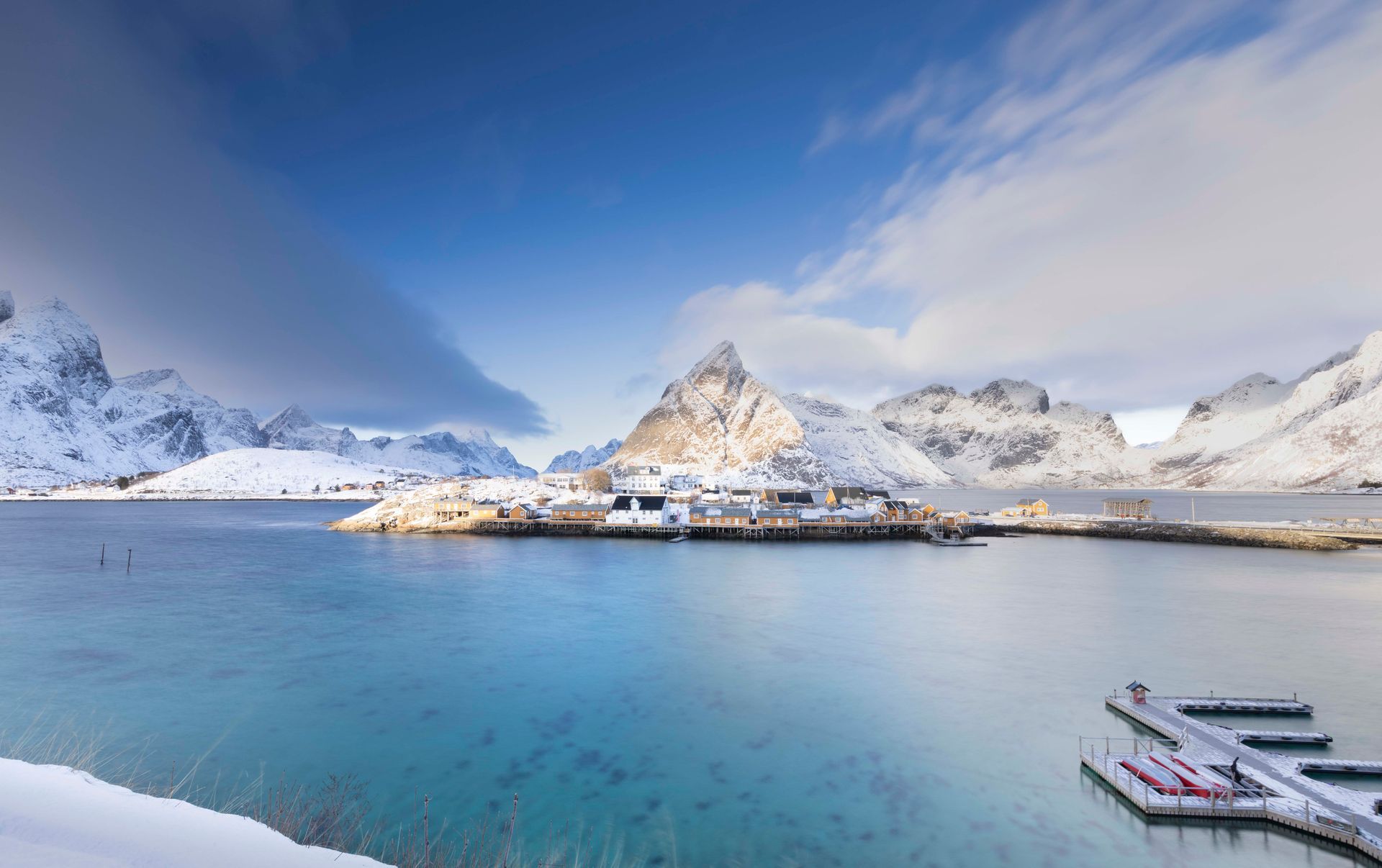 Besneeuwd bergdorp aan een turquoise fjord met aanlegsteigers op de voorgrond.