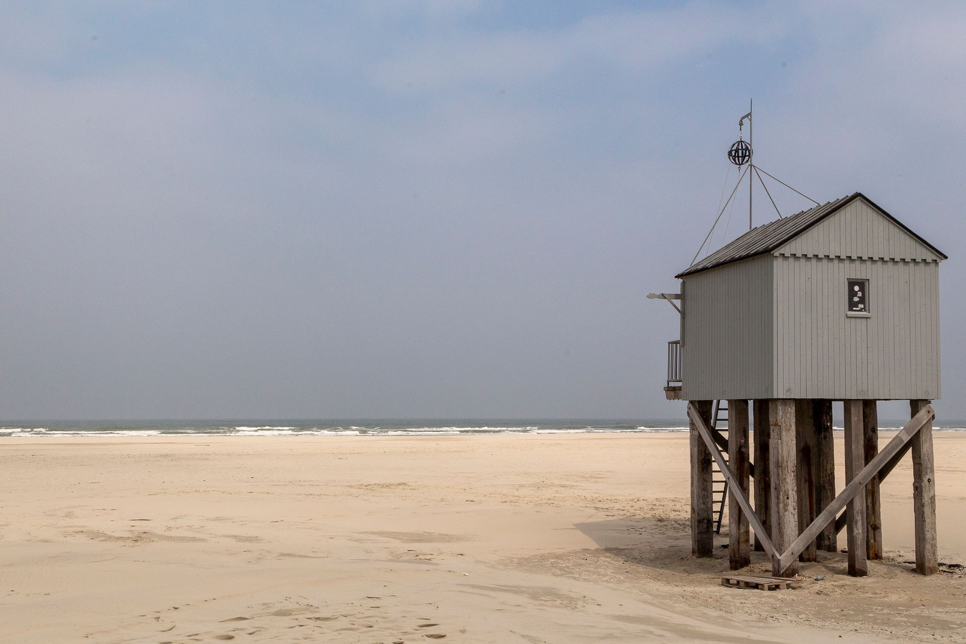Een klein grijs strandhuisje op palen naast een breed zandstrand onder een bewolkte hemel.