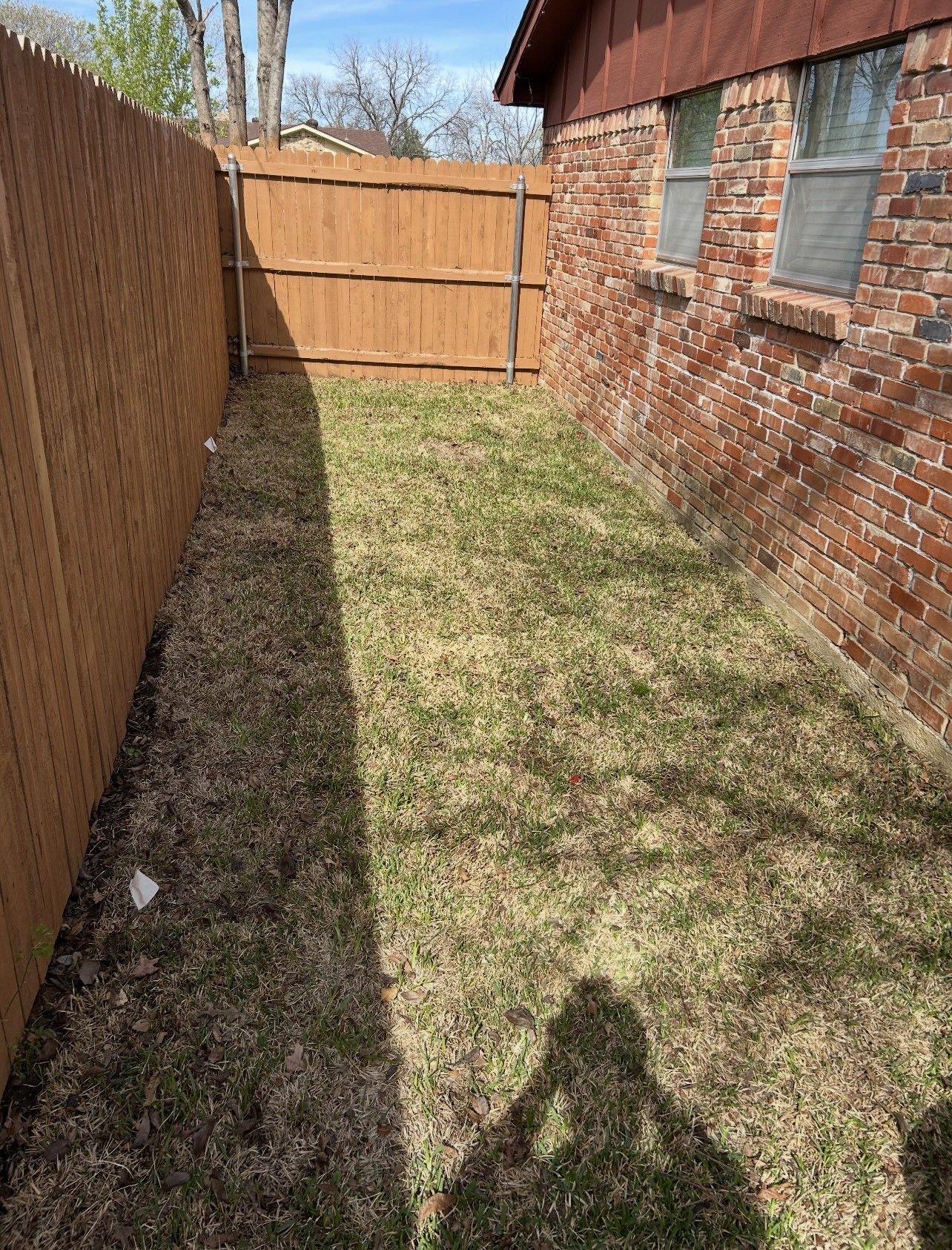 Narrow grassy side yard beside a brick house and wooden fence, ending at a closed gate.