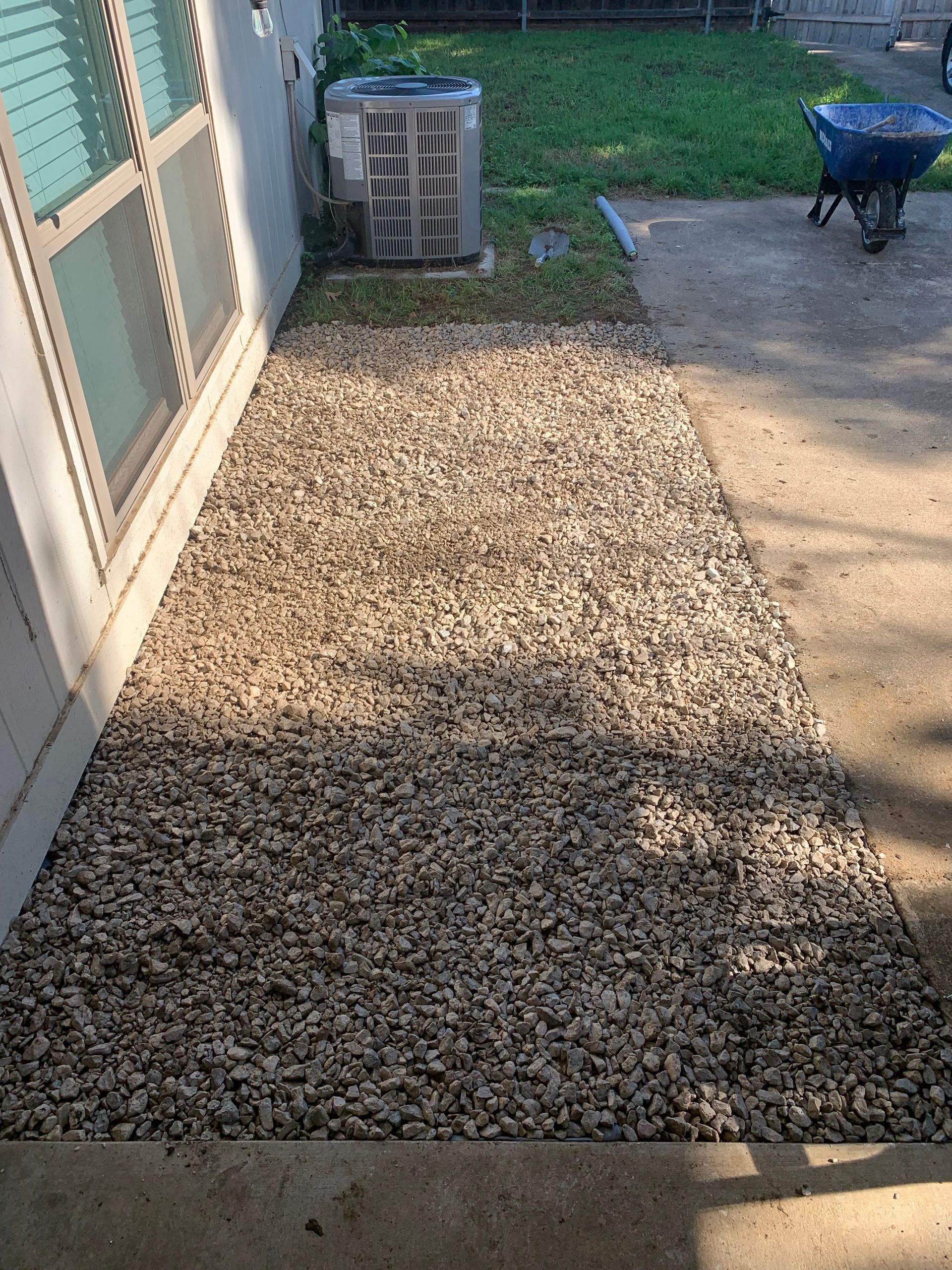 A gravel area in front of a house with an air conditioner and a wheelbarrow.