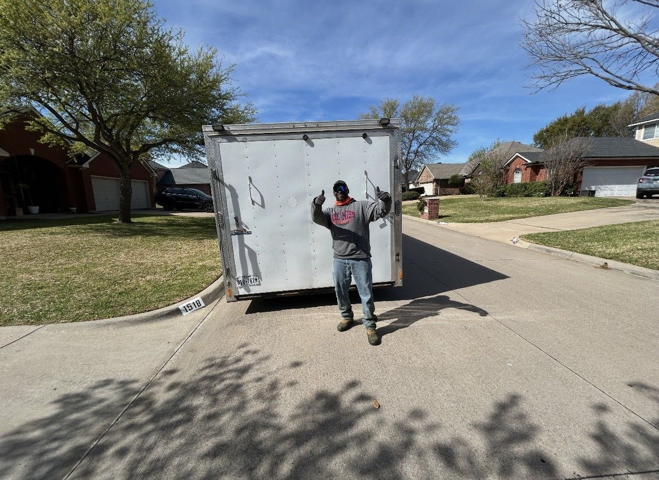 Person standing in front of a white trailer on a residential street, arms raised.