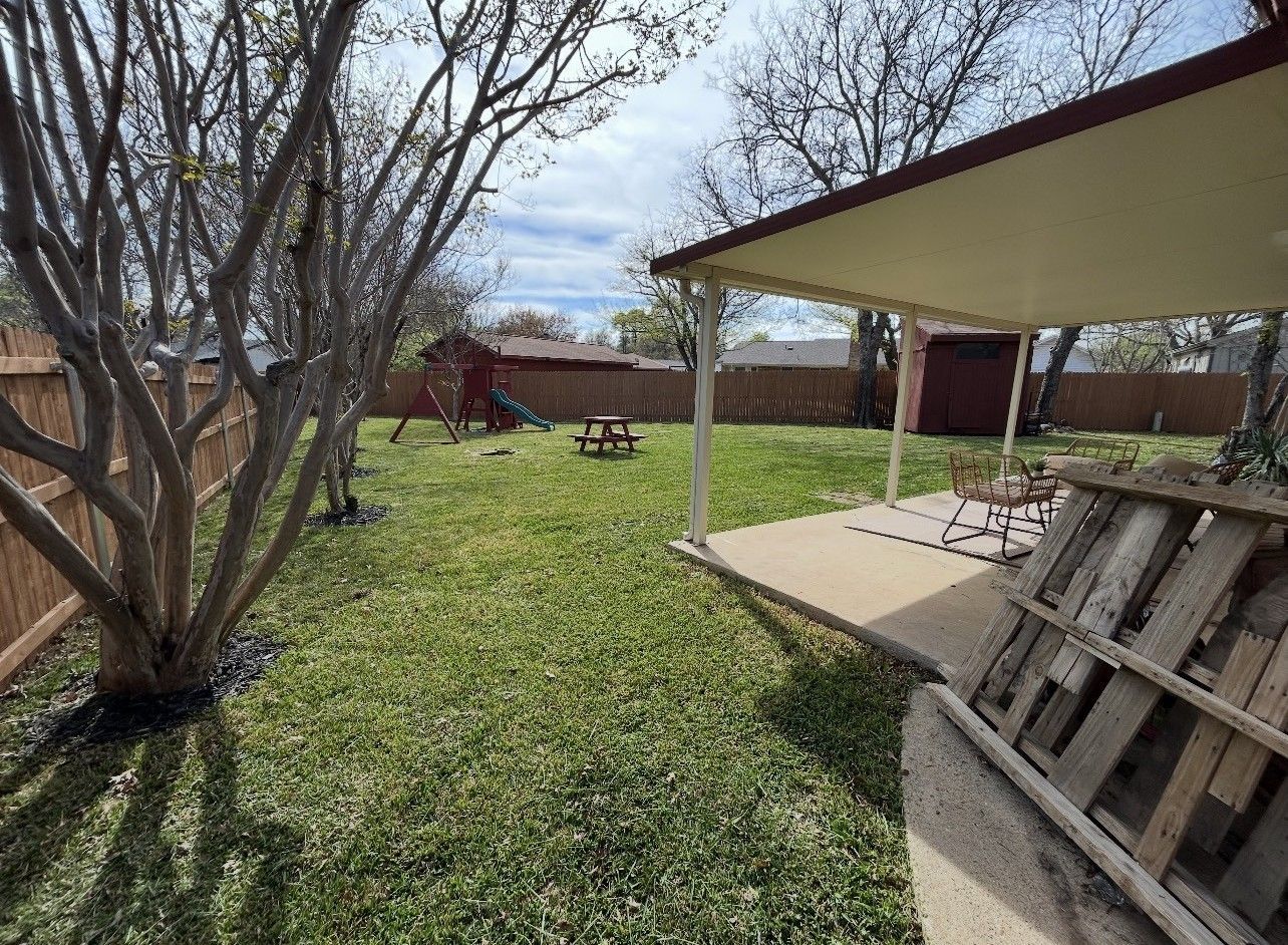 Backyard with green lawn, leafless tree, picnic table, and covered patio with stacked wood pallets