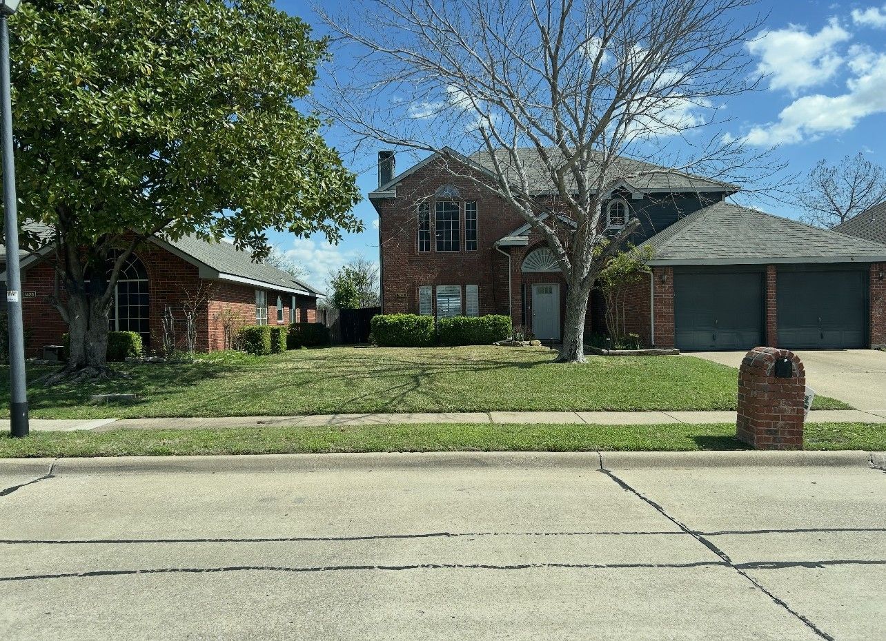 Suburban brick house with front lawn, trees, and a driveway under a blue sky