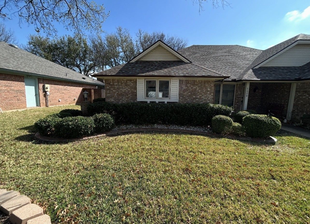Brick suburban house with a trimmed front lawn, shrubs, and a covered porch under a sunny sky