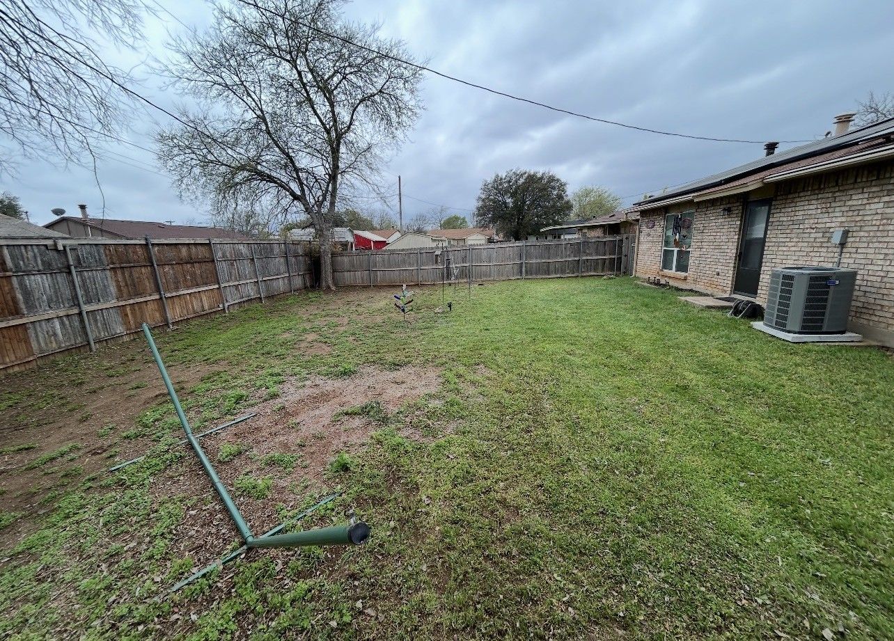 Backyard with grass, fence, bare tree, and house under cloudy sky