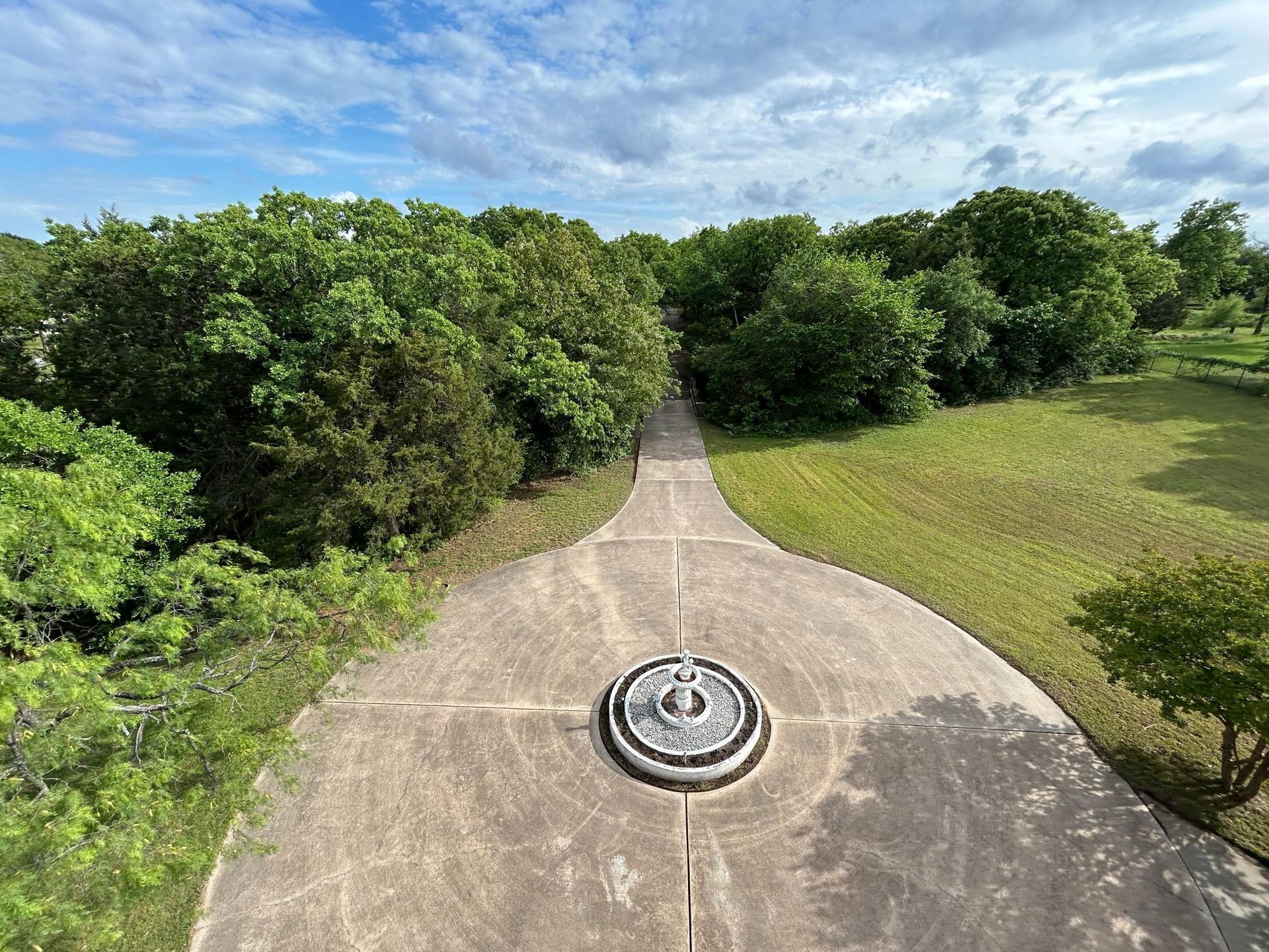 Concrete path leading through green trees to a circular fountain on a sunny day