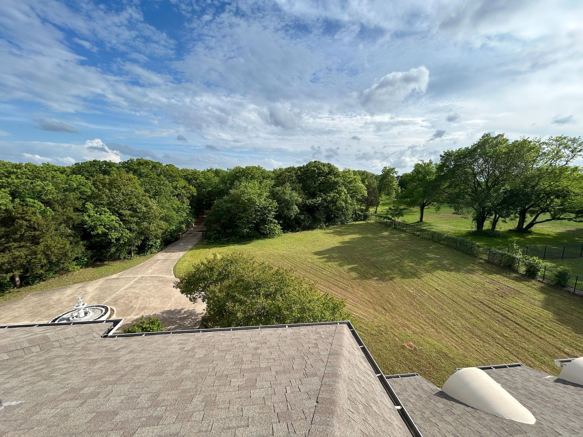 Rooftop view of a grassy yard, winding driveway, and trees under a partly cloudy sky.