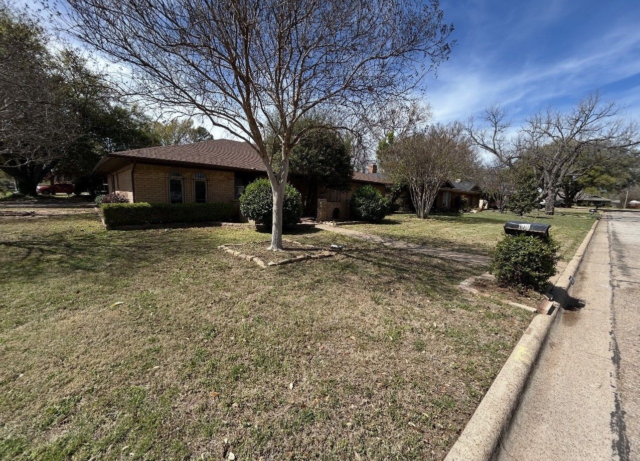 Suburban house with a large front yard, tree, and driveway beside a quiet street.