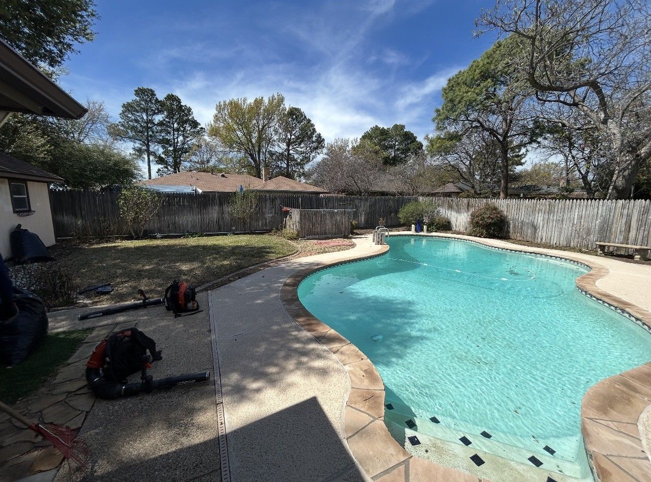 Backyard swimming pool with turquoise water, patio seating, and trees under a clear blue sky