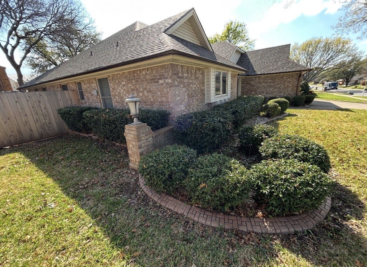 Brick house with shingled roof, fenced yard, and trimmed shrubs along a curved flower bed.