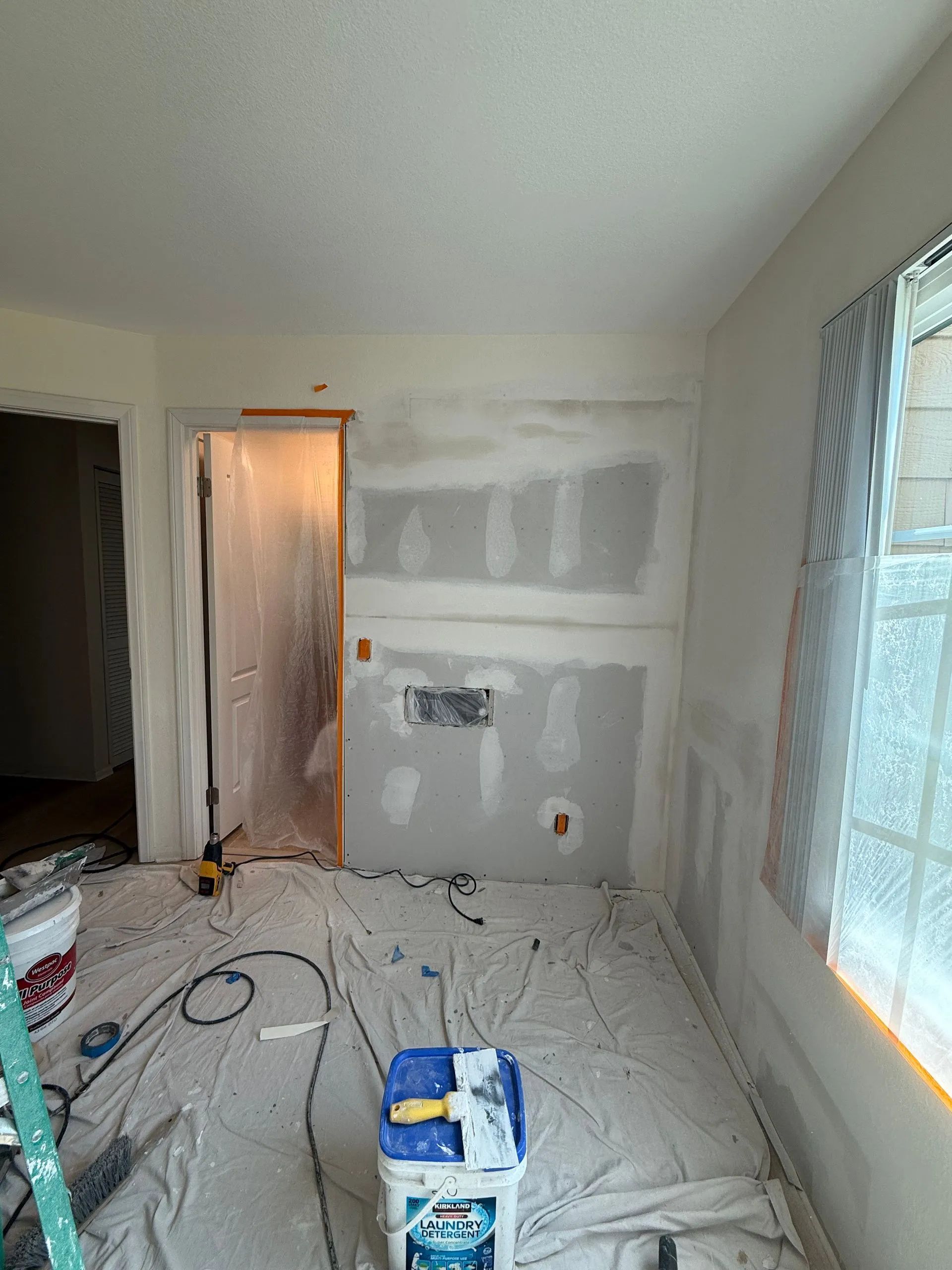 An interior room under renovation with drywall patches on the wall, drop cloths on the floor, and painting supplies.