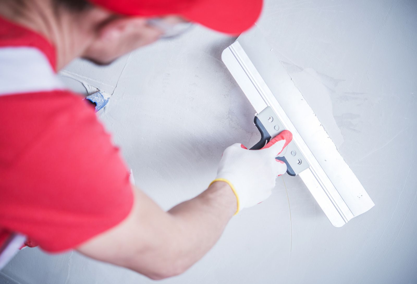 Person in red shirt using a paint roller to paint a white wall.