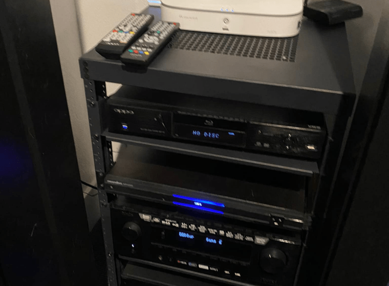 Black AV rack with stereo components and remotes on top, lit by blue display lights.