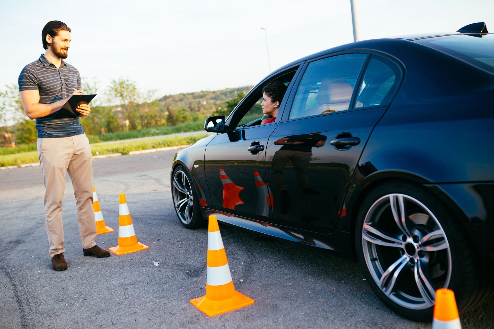 A woman is inside a black car, next to some safety cones, with a male instructor close-by. A woman is inside a black car, next to some safety cones, with a male instructor close-by.