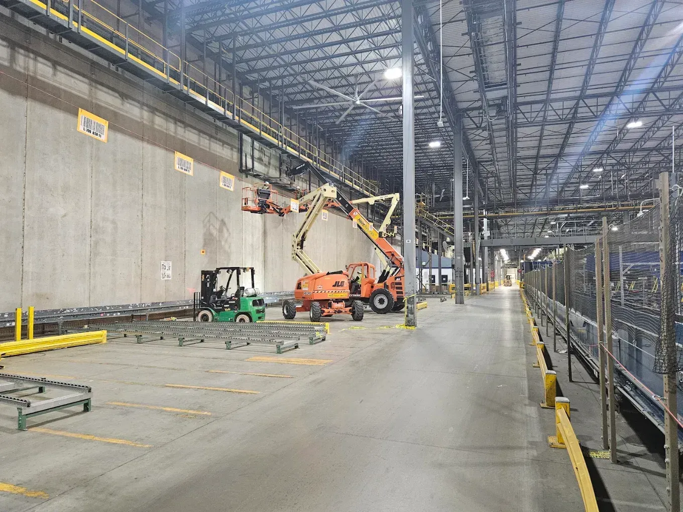 Large industrial warehouse with a lift truck and boom lift near a concrete wall under bright overhead lights