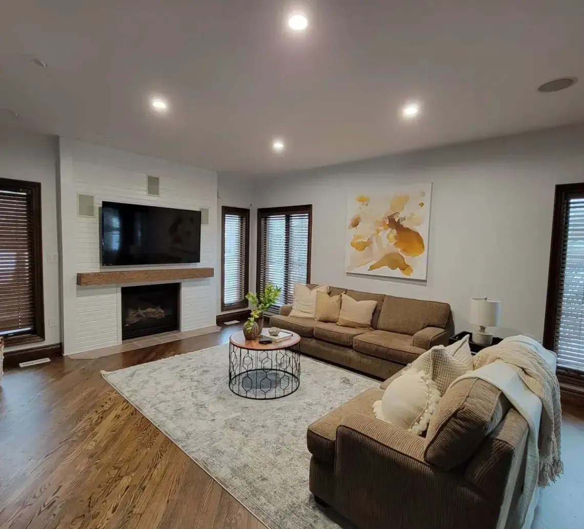 A living room with a brick fireplace, television, brown sectional sofa, area rug, and abstract art on a light wall.