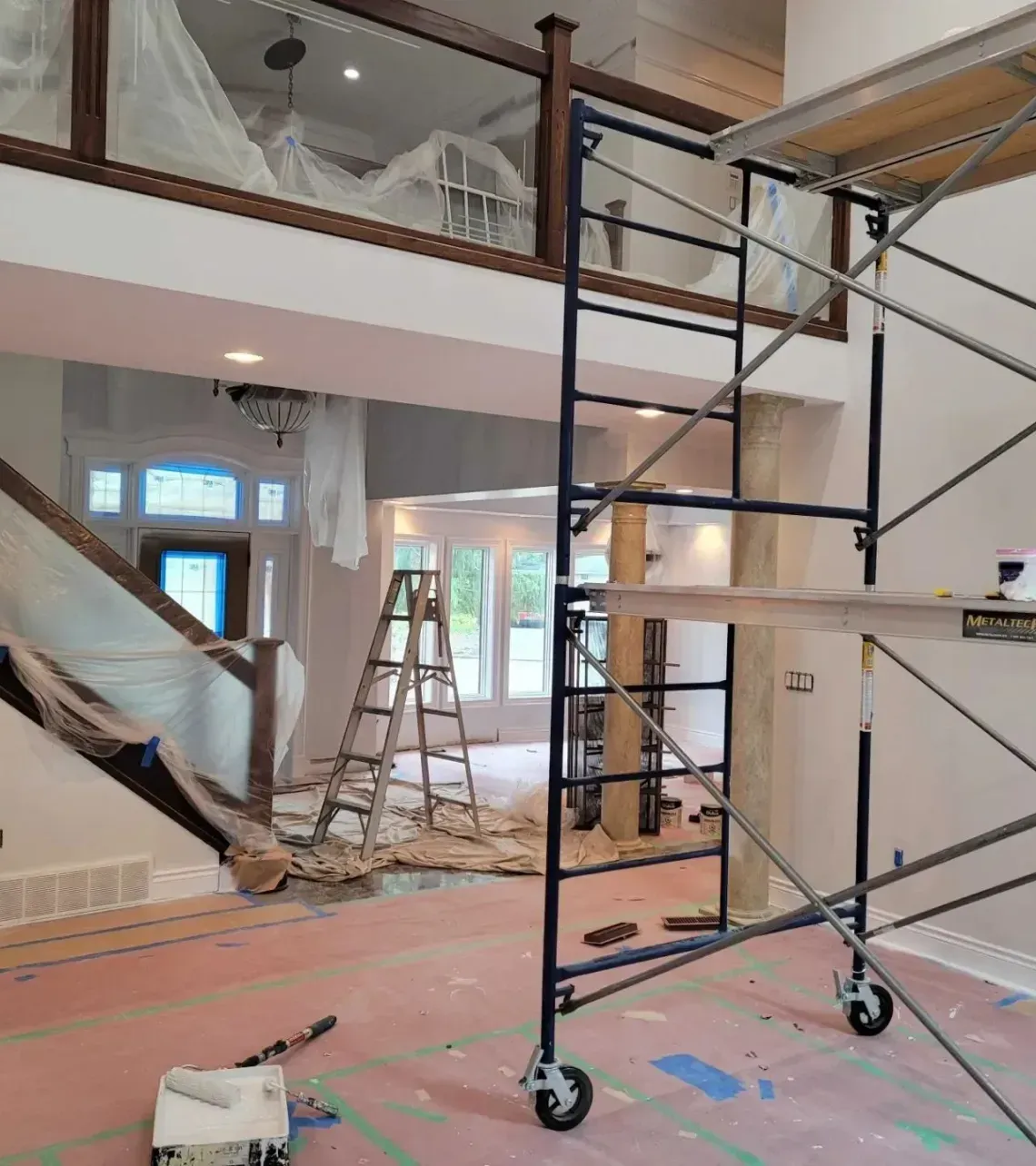 Interior of a large home under renovation, featuring a tall metal scaffold, a wooden stepladder, and plastic-covered walls.