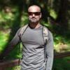 Man with sunglasses and a backpack in a forest, smiling at the camera.