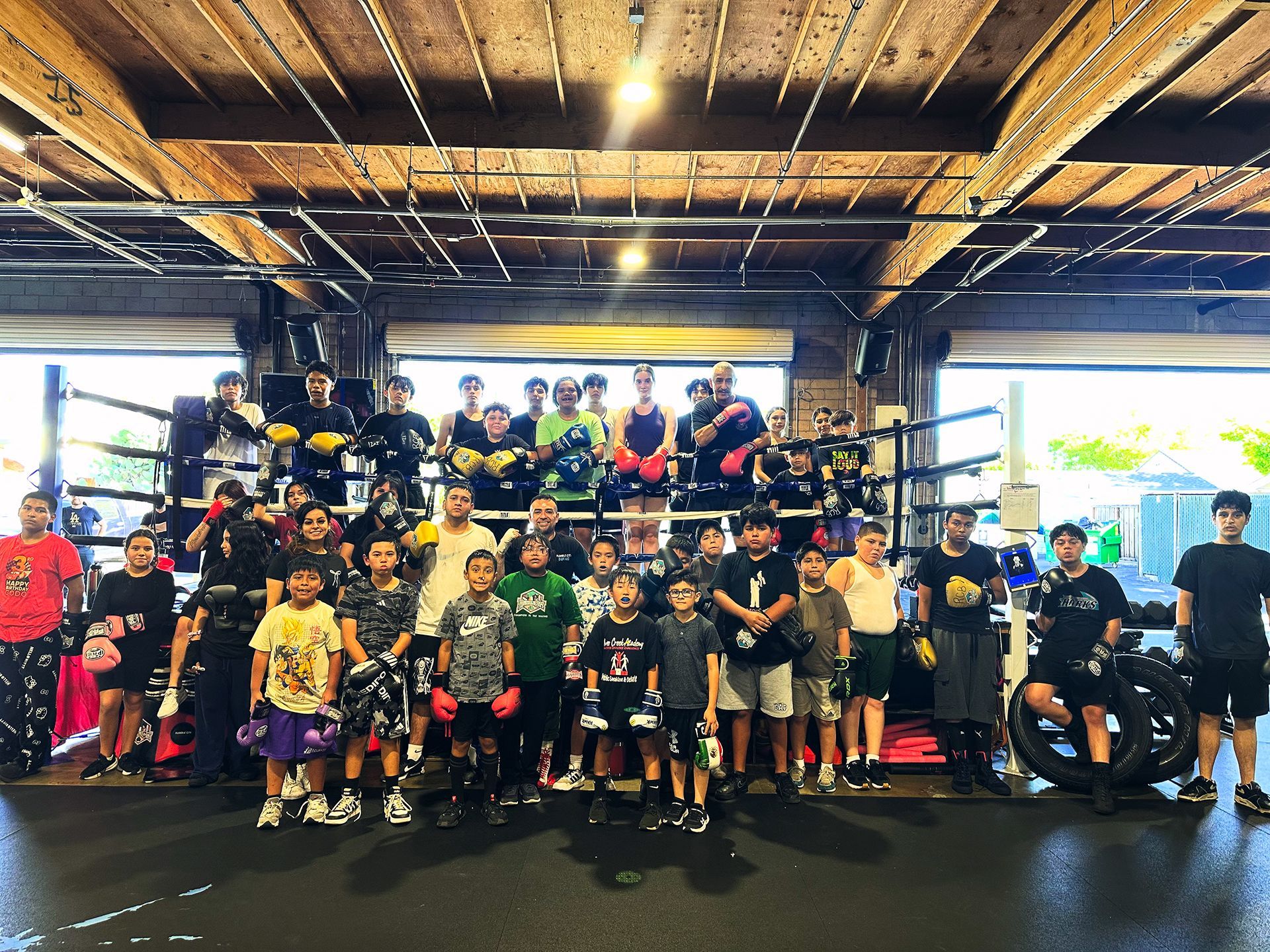 Group of boxers in a boxing ring, posing for photo. Some wear gloves, in an open-air gym.