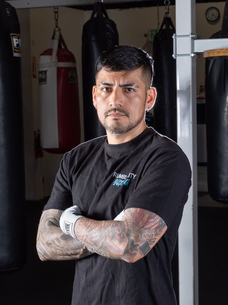 Man with crossed arms, tattoos, boxing gloves, and a serious expression in a gym with punching bags.