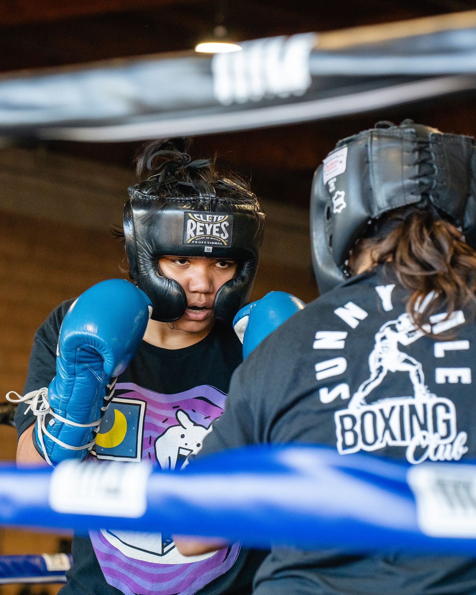 Two boxers in a ring; one with blue gloves and headgear, facing the other, both preparing to spar.