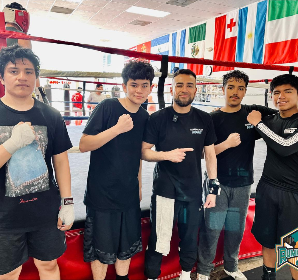 Five young men in a boxing ring with flags. They pose with fists up. One man points.