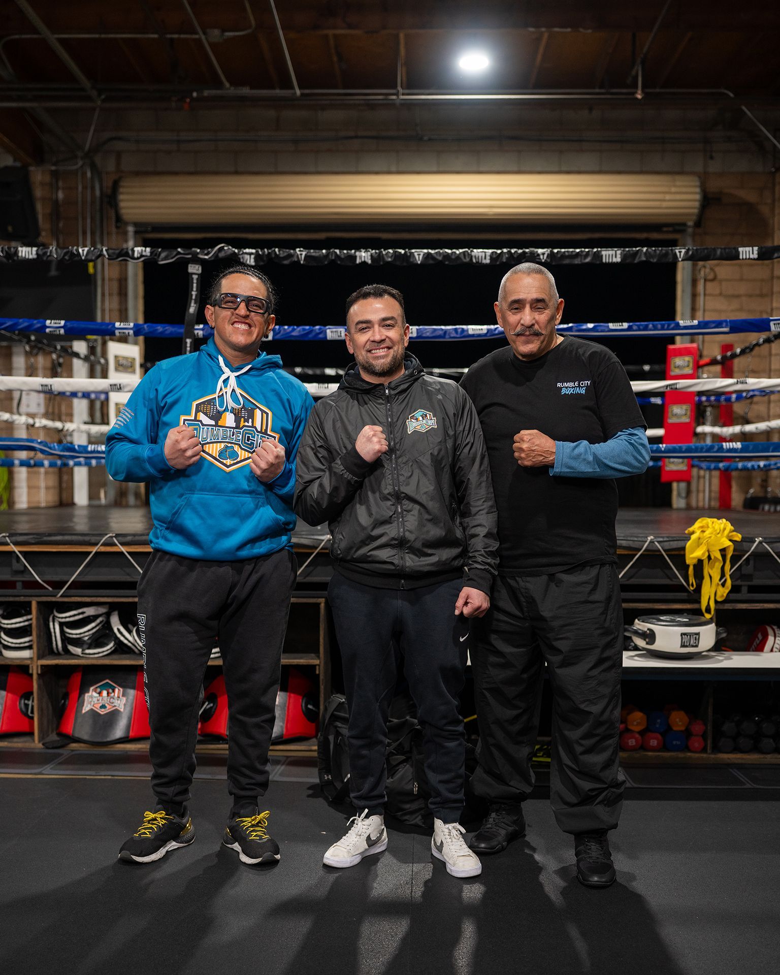 Three men in a boxing ring, posing. Two are smiling. One wears a blue hoodie, the others wear jackets.