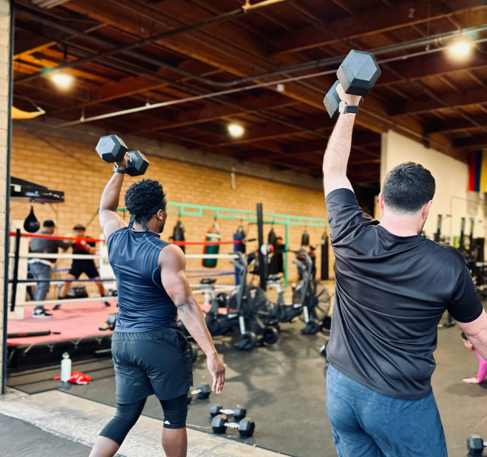 Two men lifting dumbbells overhead in a gym, with boxing ring and workout equipment visible in background.