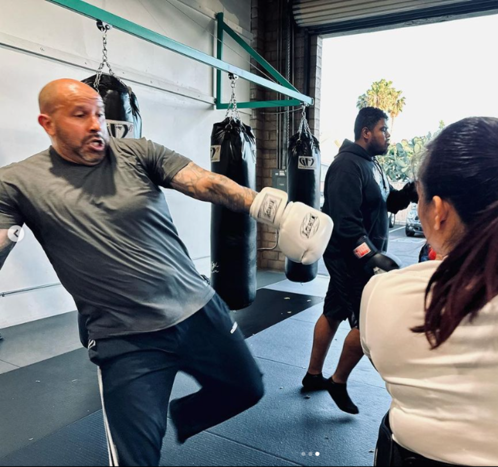 Man in a boxing stance, throwing a punch at a woman. Training in a gym, other people in background.