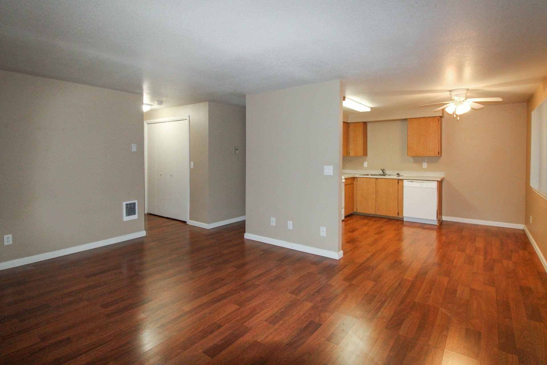 An empty living room with hardwood floors and a kitchen in the background.