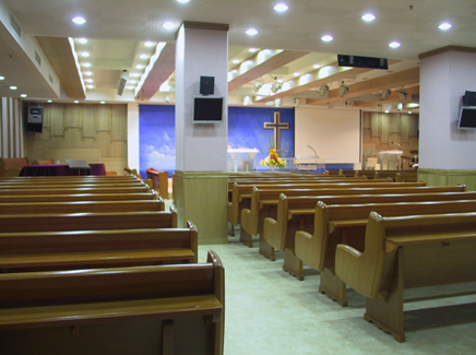 Compact church setup with wooden benches, stage cross, and simple sound system.