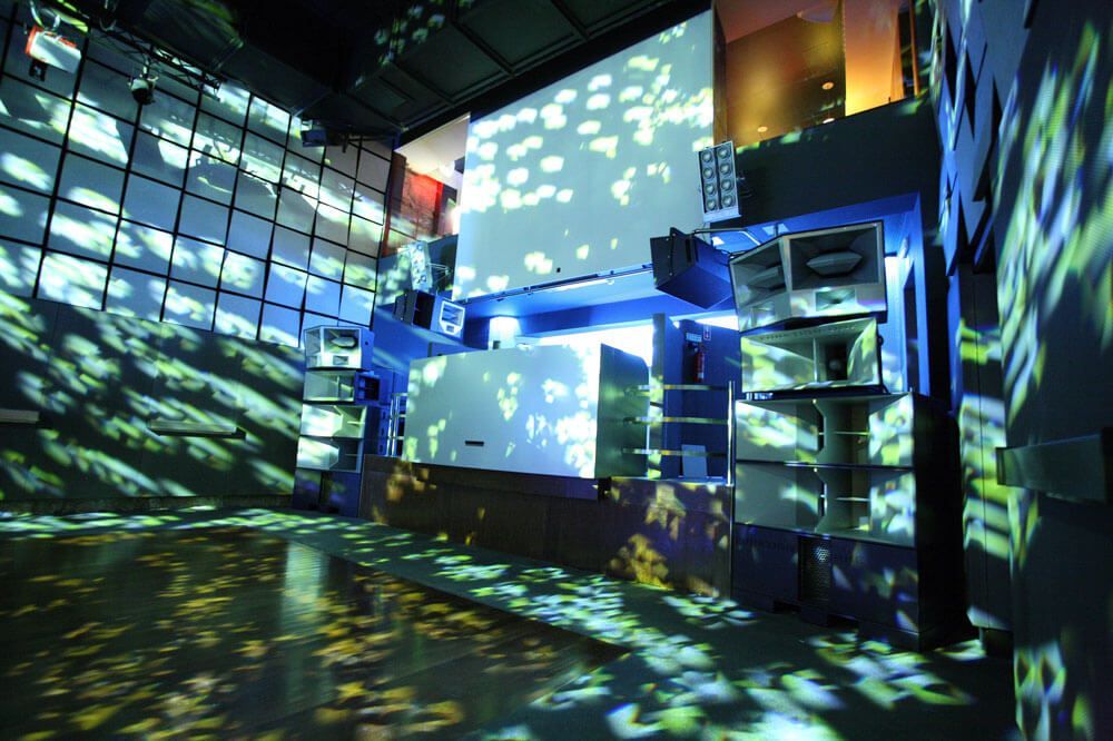 Nightclub interior with a powerful sound system, large speakers, and colorful light projections, creating a vibrant atmosphere.