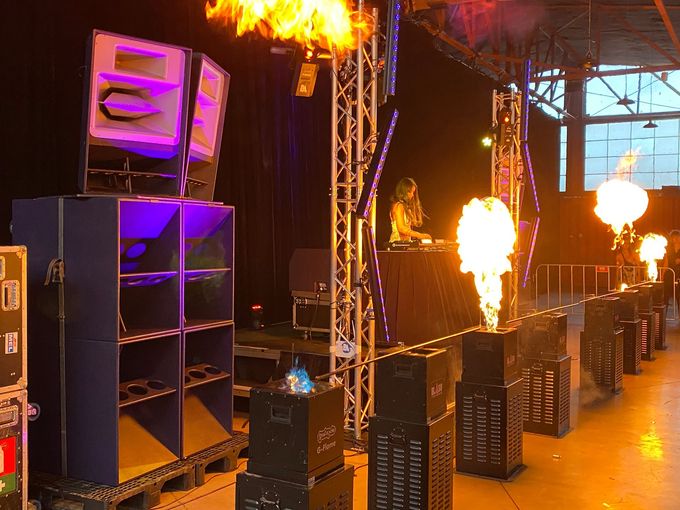 DJ performing live on stage with large speakers, LED lighting, and pyrotechnic flame effects at an indoor music event.