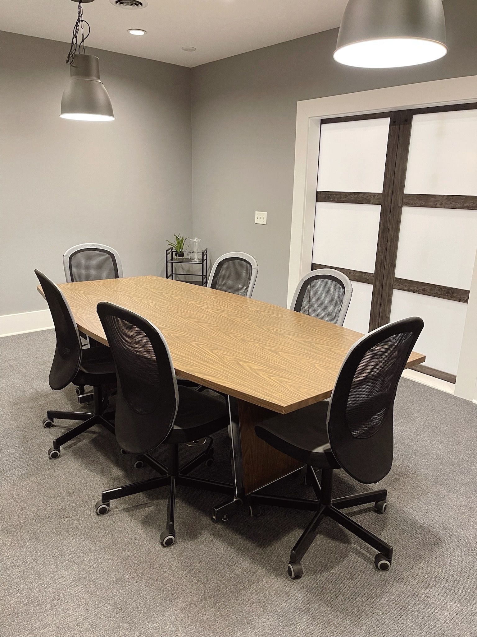 Conference room with long wooden table, black chairs, pendant lights, and frosted glass doors.