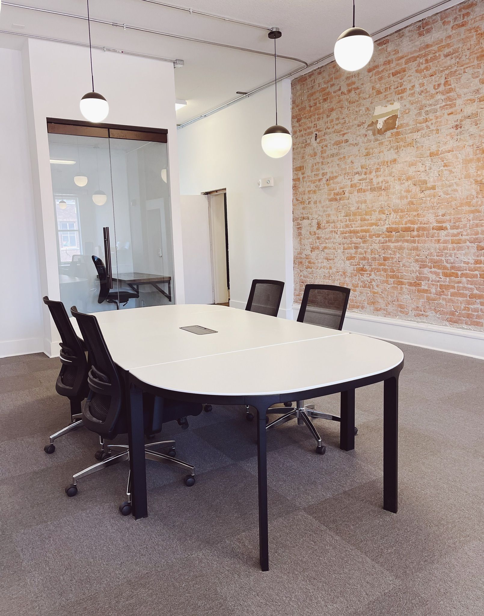 Modern conference room with white oval table, black chairs, hanging lights, and exposed brick wall