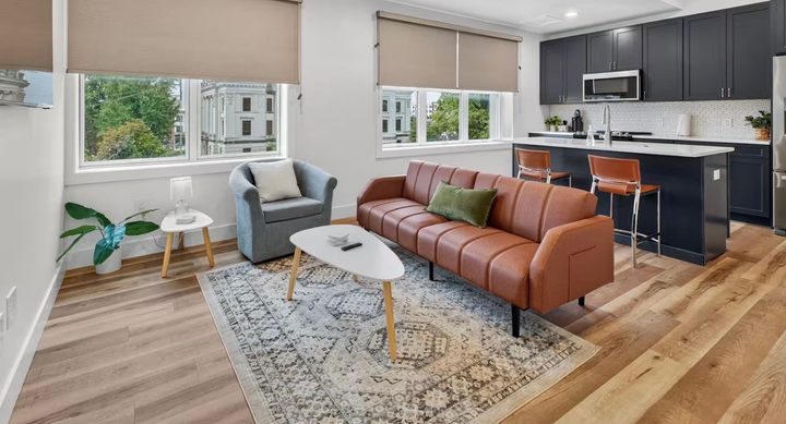 Modern living room with brown sofa, gray chair, patterned rug, and open kitchen with dark cabinets