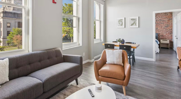 Bright modern living room with gray sofa, tan chair, dining table, and large windows