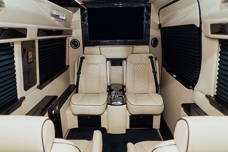 Interior of a luxury van with cream-colored leather seating, a large screen, and dark window shades.
