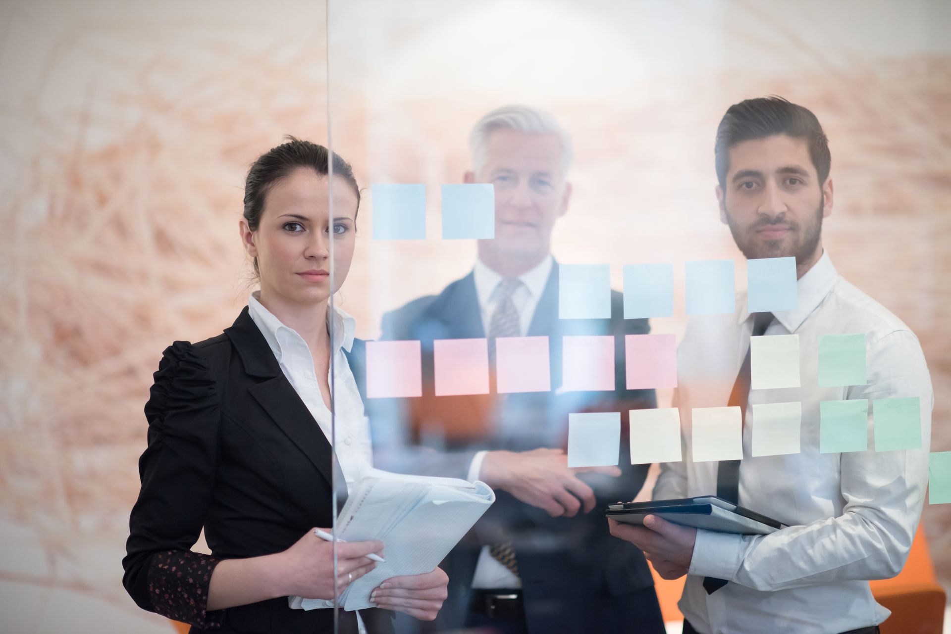 Een zakelijk team van drie personen bekijkt post-it-briefjes op een glazen wand.