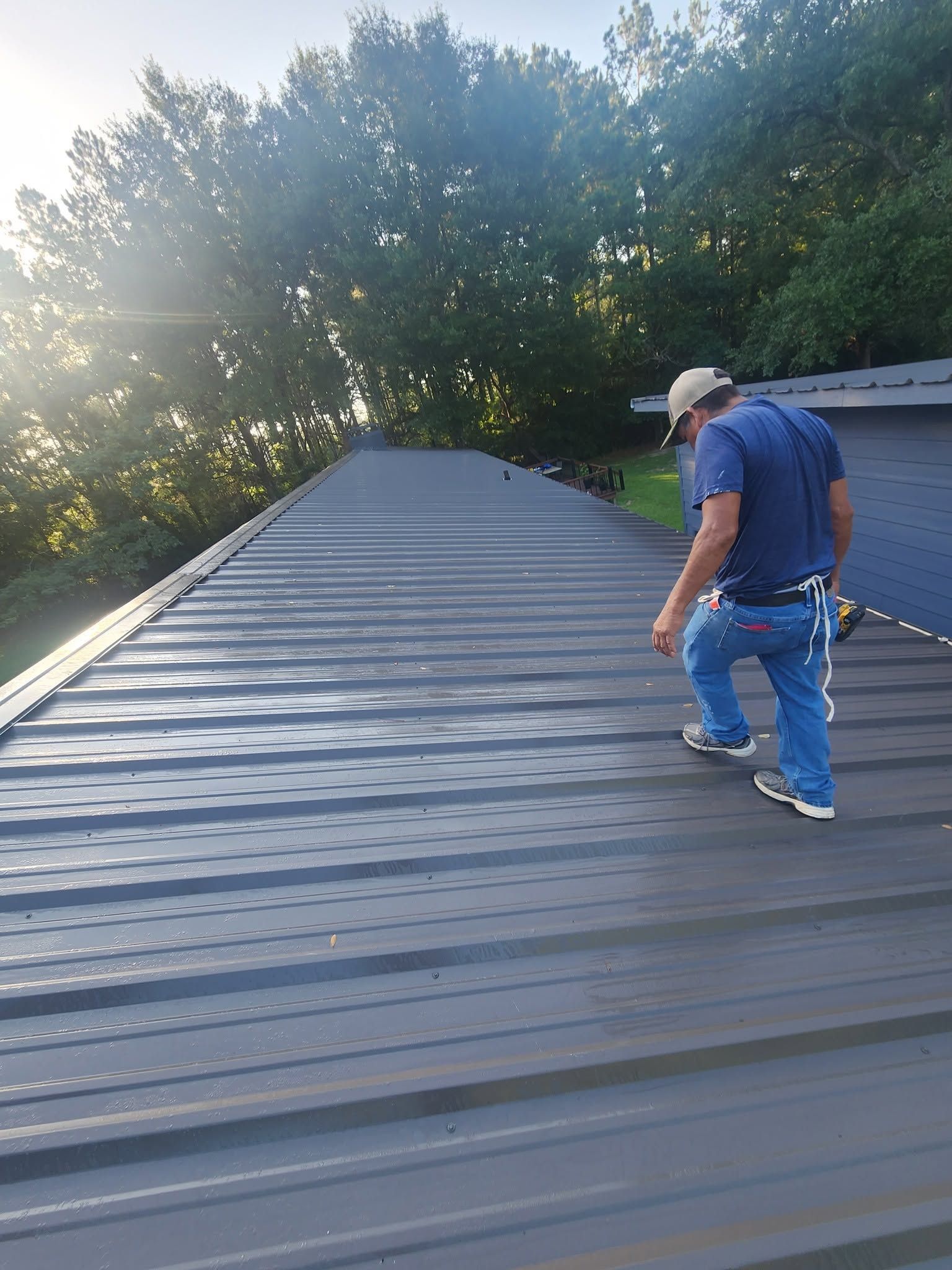 Worker inspecting a gray metal roof in sunlight, with trees in the background.