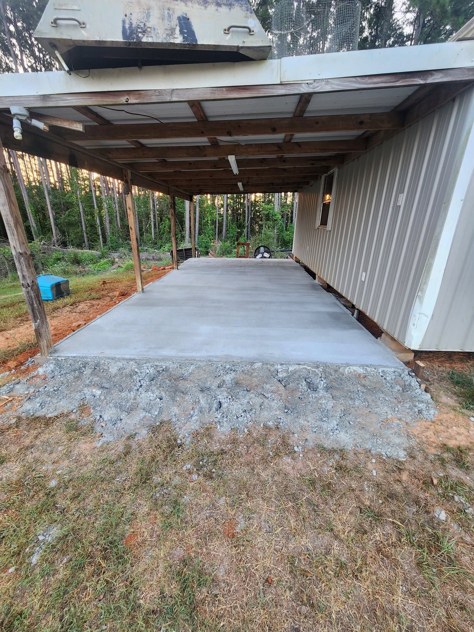 New concrete slab beside a corrugated metal building with a low carport roof and wooded yard.
