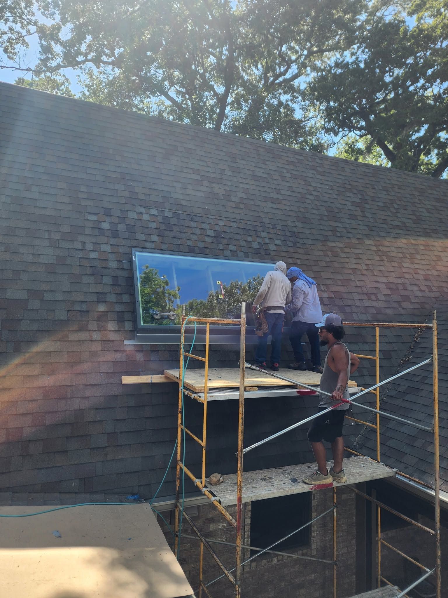 Workers on scaffolding painting a brick wall beside a roofline under trees.